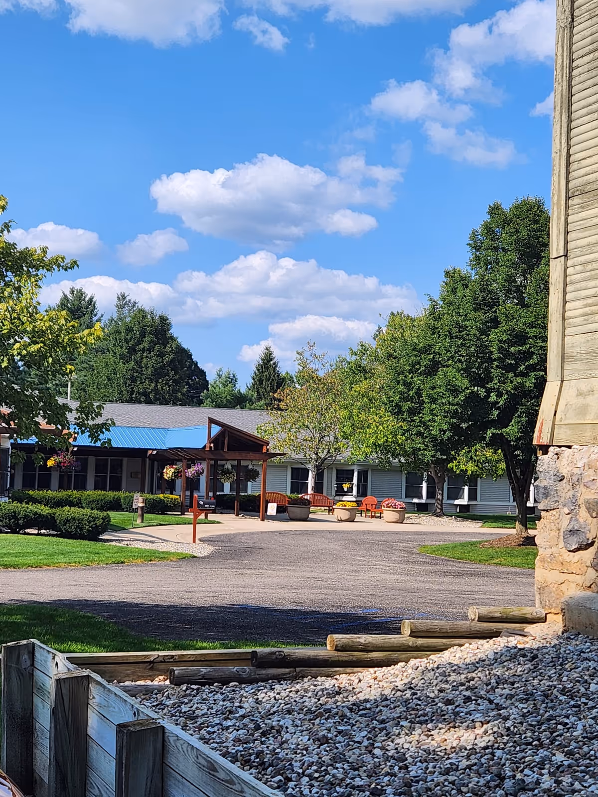 Front exterior of a senior living building with an entry canopy, benches, planters, trees and a driveway under a blue sky.