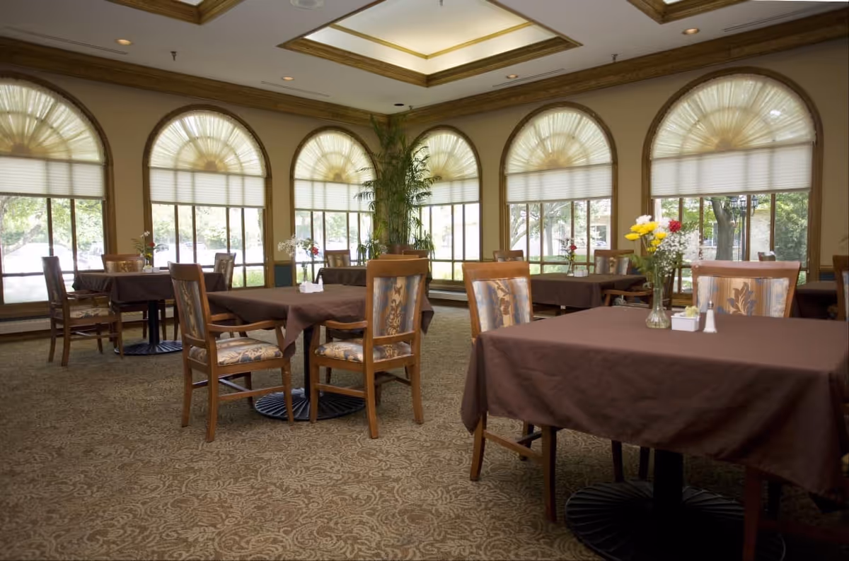 Dining room with multiple tables covered in brown tablecloths, wooden chairs, arched windows with shades, and floral centerpieces.