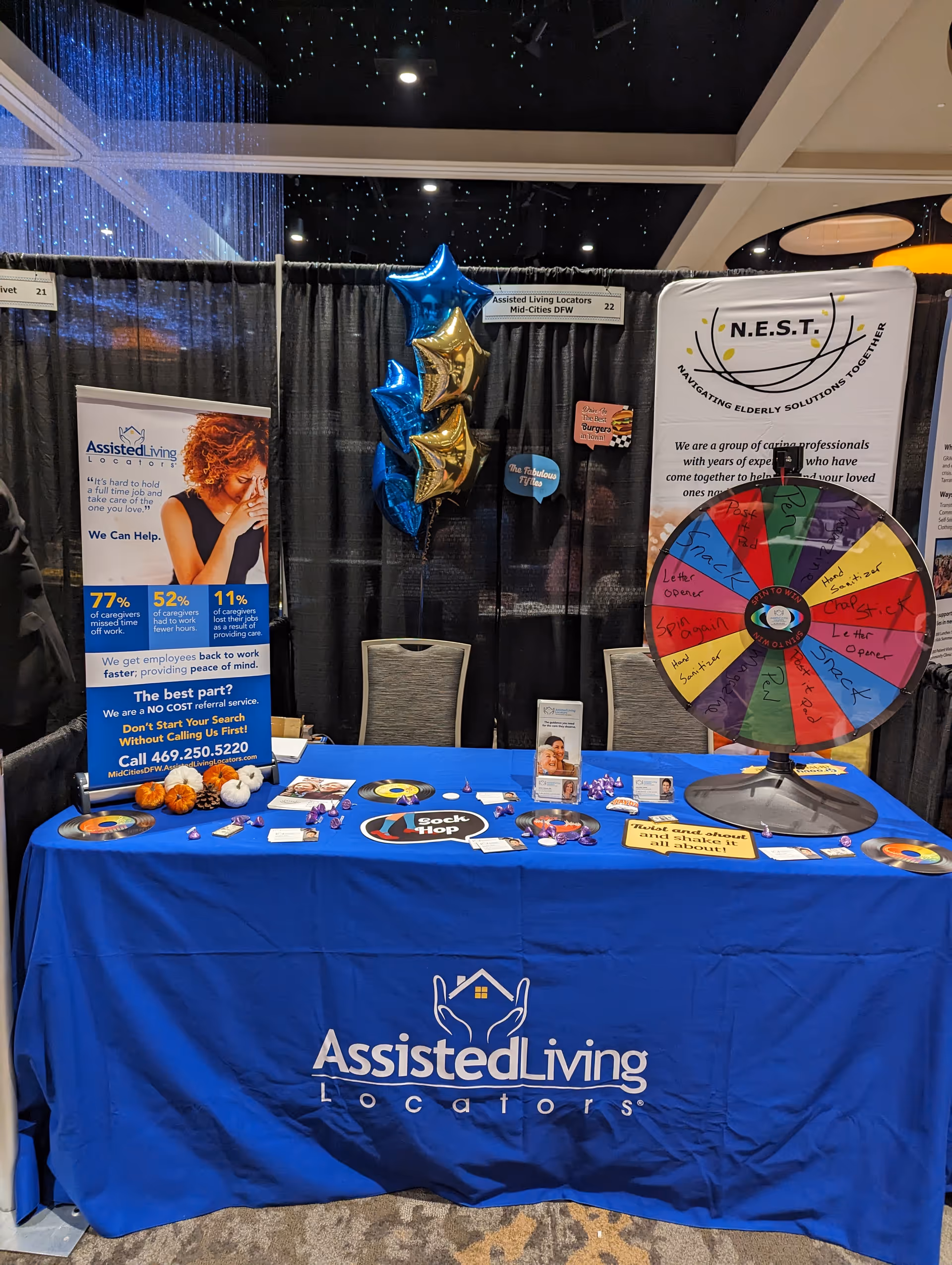 Blue tablecloth expo booth for Assisted Living Locators featuring a wheel of fortune and banners.