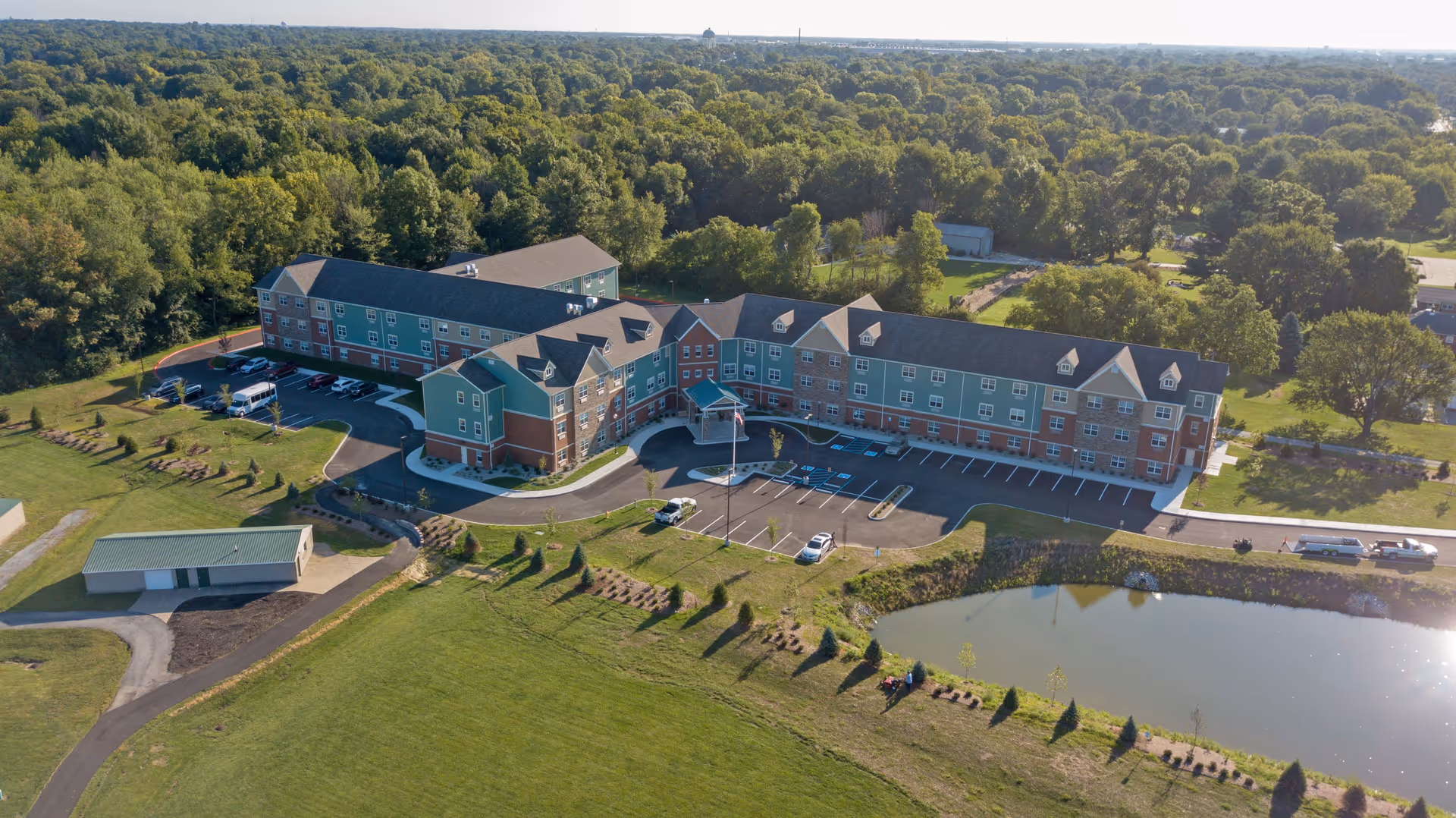 Aerial view of Glasswater Creek of Lafayette, a large senior living facility surrounded by greenery and trees. The building is multi-story with a mix of brick and green siding, featuring multiple windows and a covered entrance. There is a parking lot with several cars, a small pond in the foreground, and landscaped grounds with pathways and small trees.