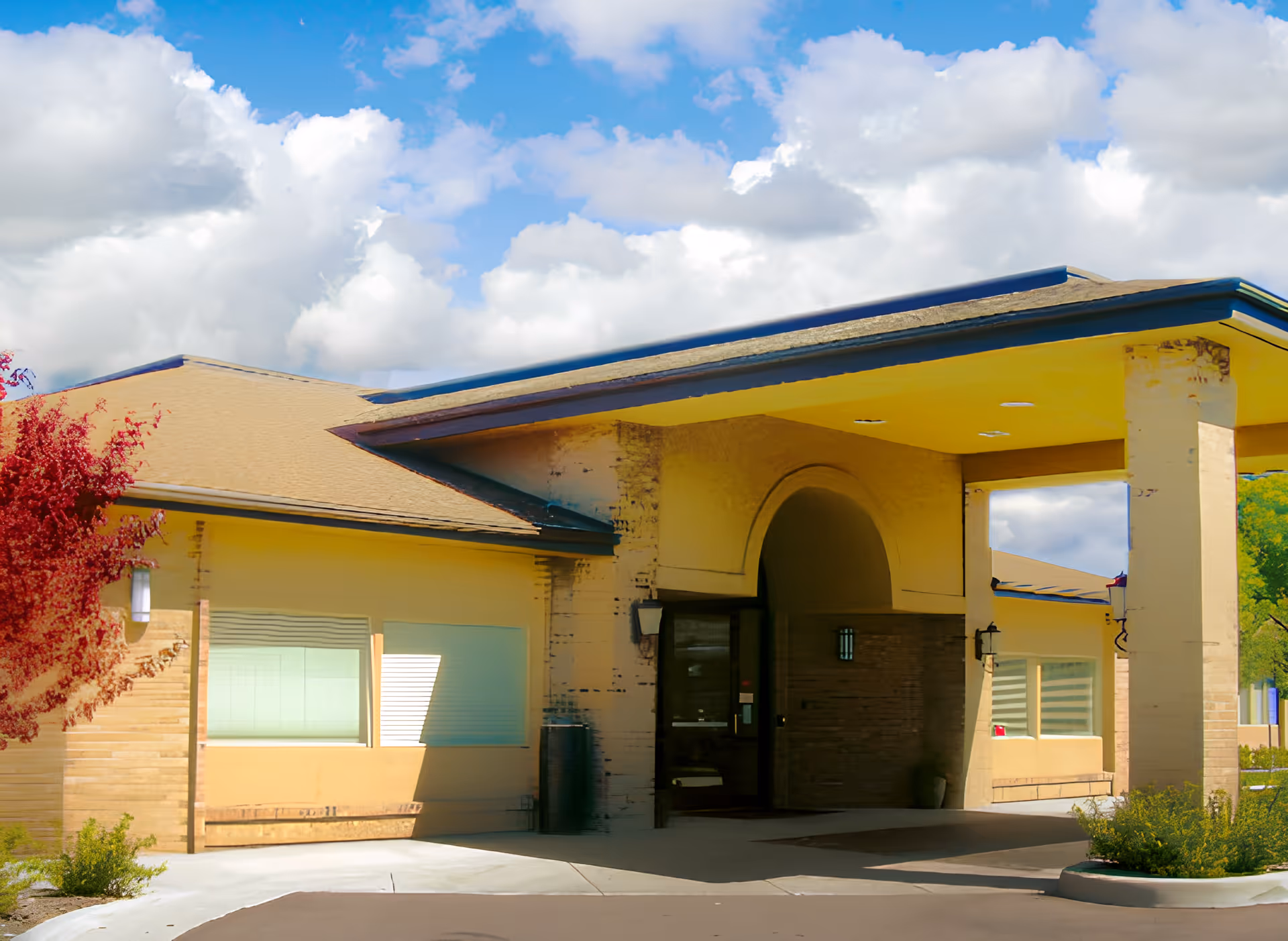 Exterior view of a single-story building with a covered entrance supported by columns. The building has beige walls, a sloped roof, and large windows with blinds. There is a small landscaped area with bushes and a tree with red leaves near the entrance. The sky is partly cloudy with blue patches visible.