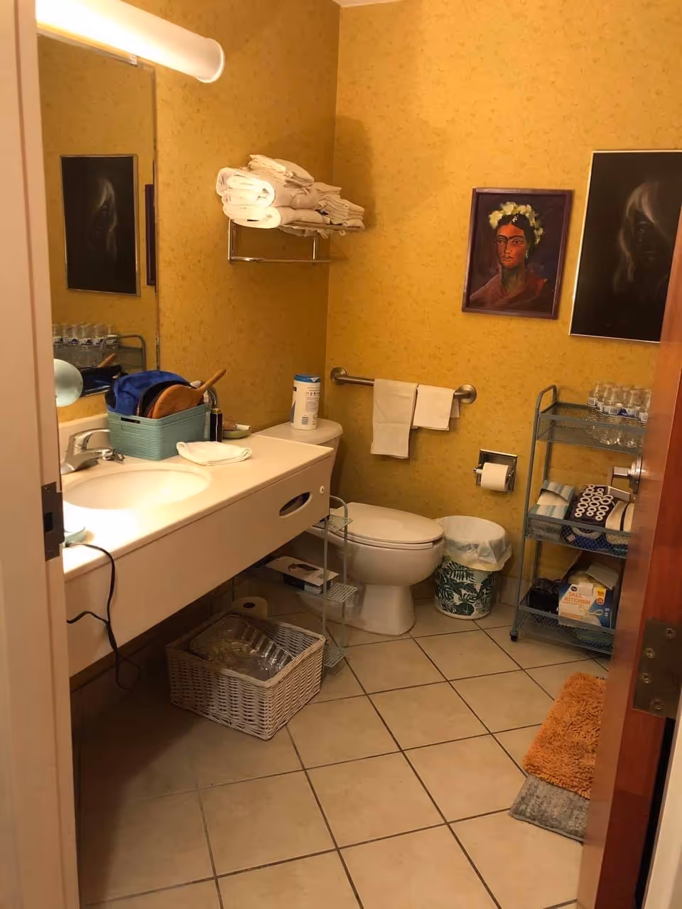 Small bathroom with a sink and countertop on the left, a toilet and metal shelving holding towels and toiletries against yellow wallpaper and a tiled floor.