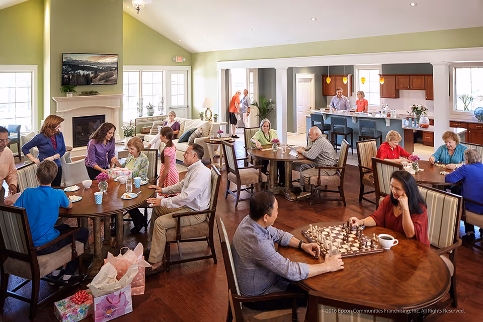 A bright and spacious common area in a senior living facility with multiple round wooden tables where elderly residents and visitors are engaged in various activities such as playing chess, eating, and socializing. The room features large windows, a fireplace with a mounted TV above it, and an open kitchen area in the background with two people standing behind the counter. The atmosphere is warm and lively with natural light filling the space.