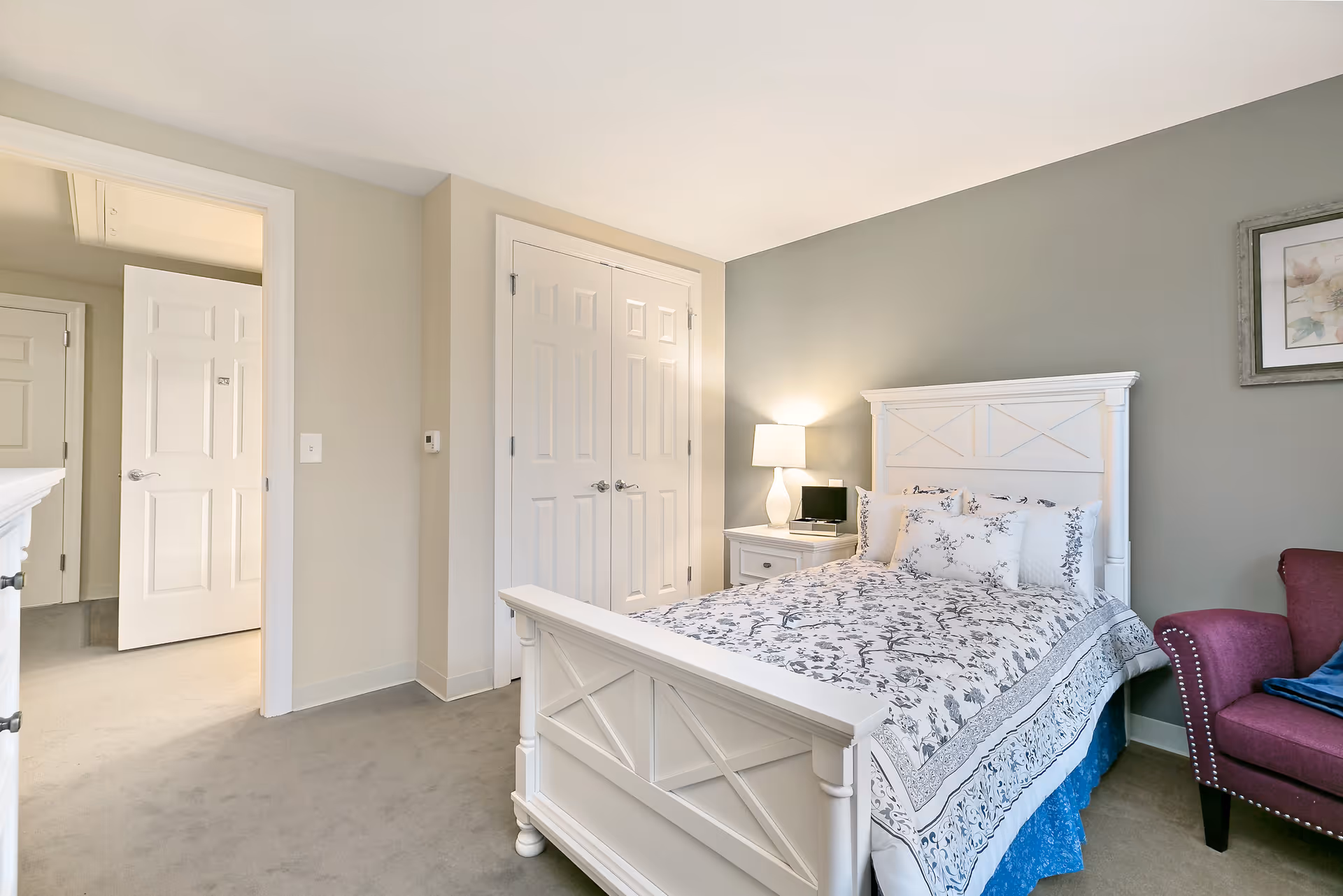 A bright bedroom with a white wooden bed frame and floral patterned bedding. There is a white nightstand with a lamp and a small digital clock or device on it. A maroon upholstered chair with a blue cushion is positioned near the bed. The walls are painted in a soft green and beige color, and there is a framed picture hanging on the wall. The room has beige carpeting and white doors, including a closet with double doors and an open door leading to another room.