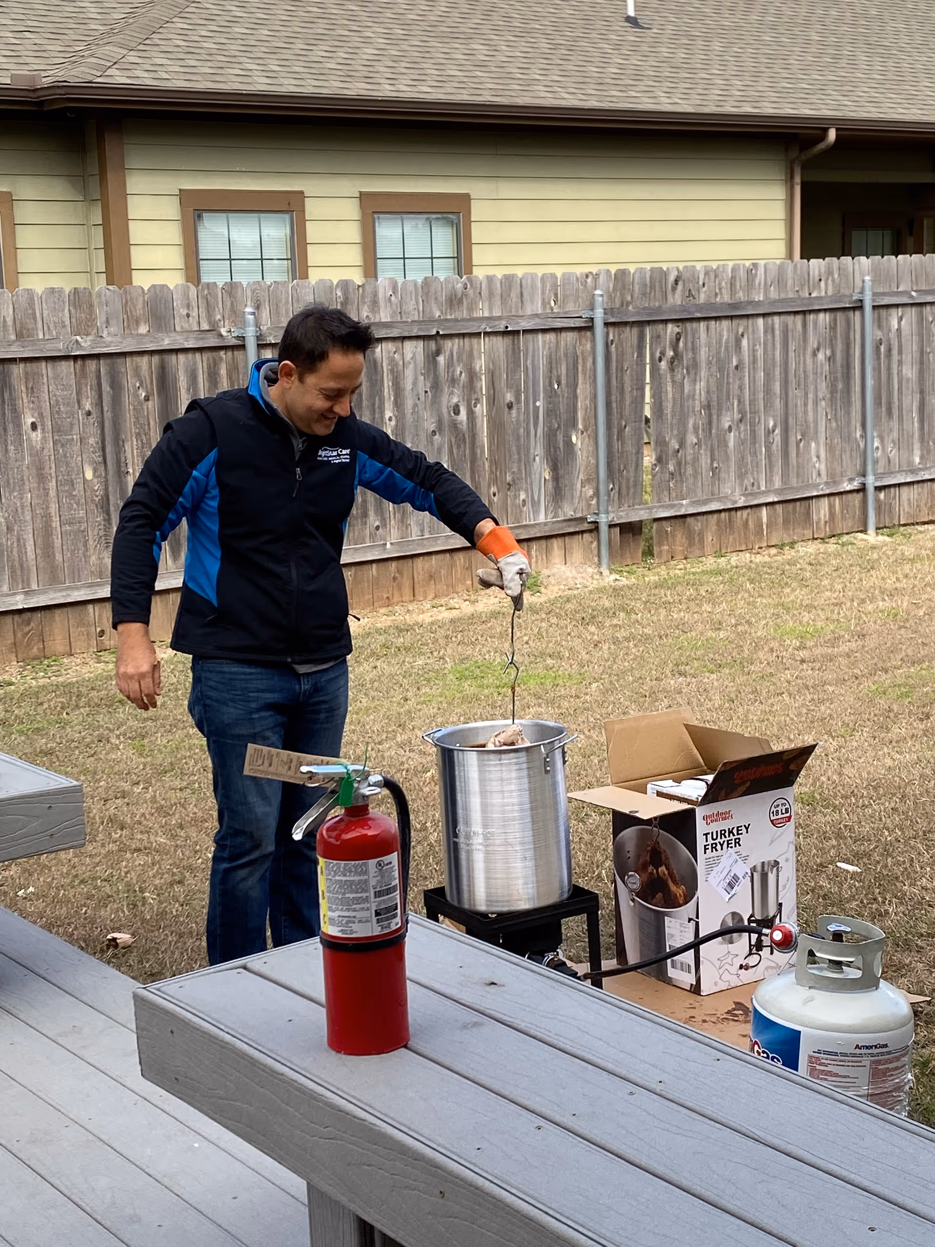 A man in a dark jacket prepares food outdoors with a turkey fryer and propane tank.