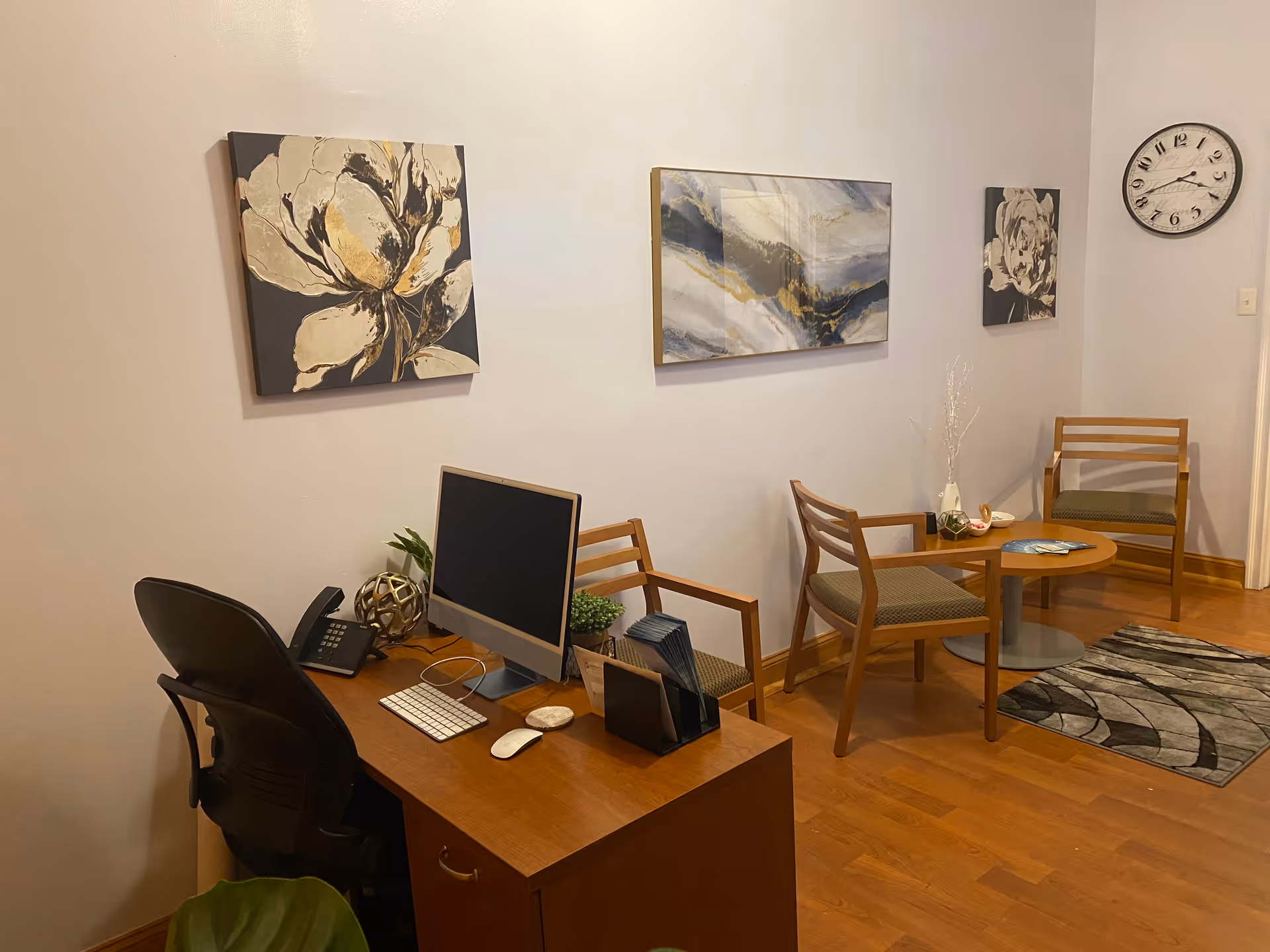 Office-like reception lounge with desk, computer, chairs, wall art, and a clock.