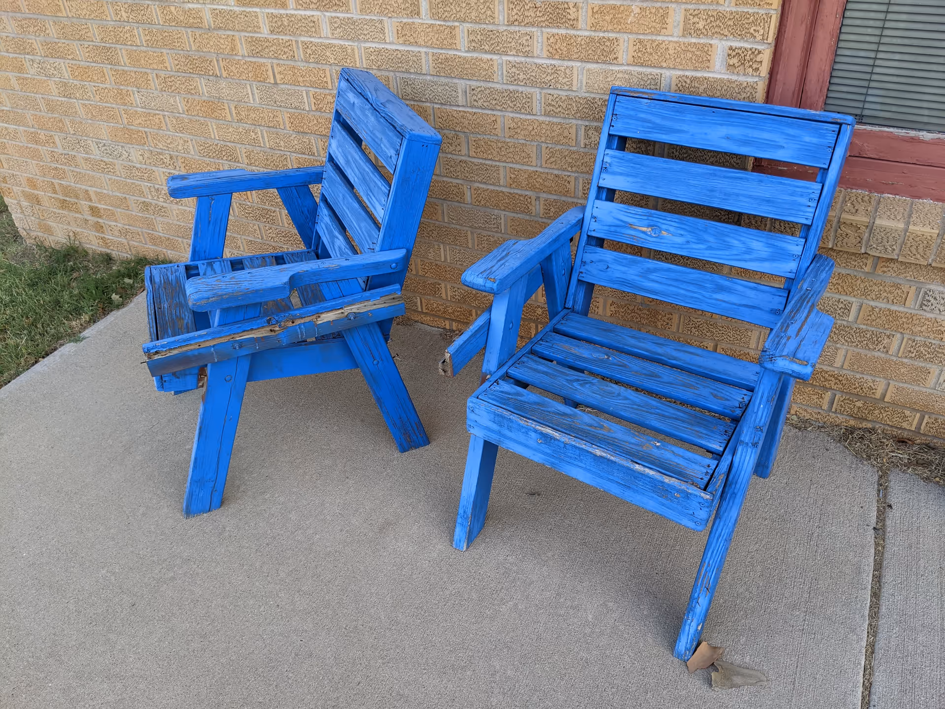 Two blue wooden chairs on a concrete patio beside a brick building.