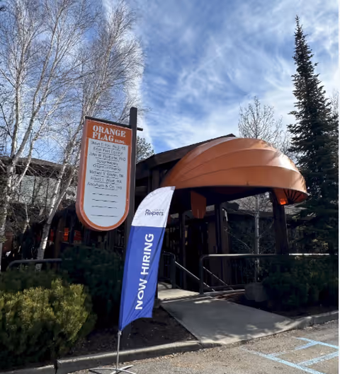 Facility entrance with an orange curved canopy, ramp, trees, and a now hiring banner.