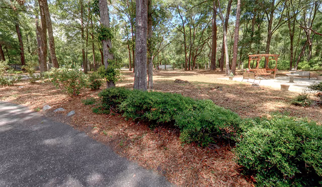 Shaded outdoor area with trees, shrubs, mulch, and a wooden bench on a paved path.