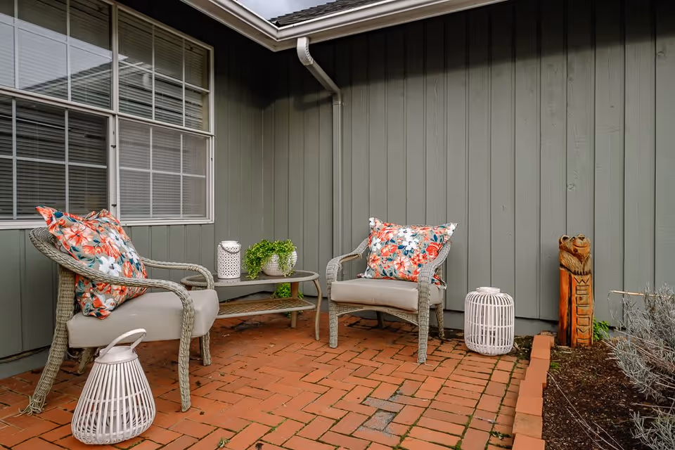 Outdoor patio area with two wicker chairs featuring floral cushions, a small glass-top table with a potted plant and decorative lanterns, set against a green wooden wall with a window and a carved wooden bear statue.