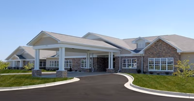 Front exterior of a single-story senior living facility with a covered porte-cochere entrance, stone siding, and curved driveway.