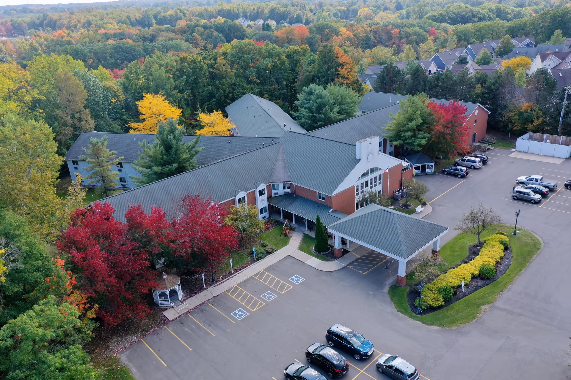Aerial view of a senior living facility surrounded by trees with autumn foliage. The building has a large covered entrance with a driveway and parking spaces, including several handicap spots. There are cars parked in the lot and a small gazebo near the building. The facility is located in a suburban area with houses visible in the background.