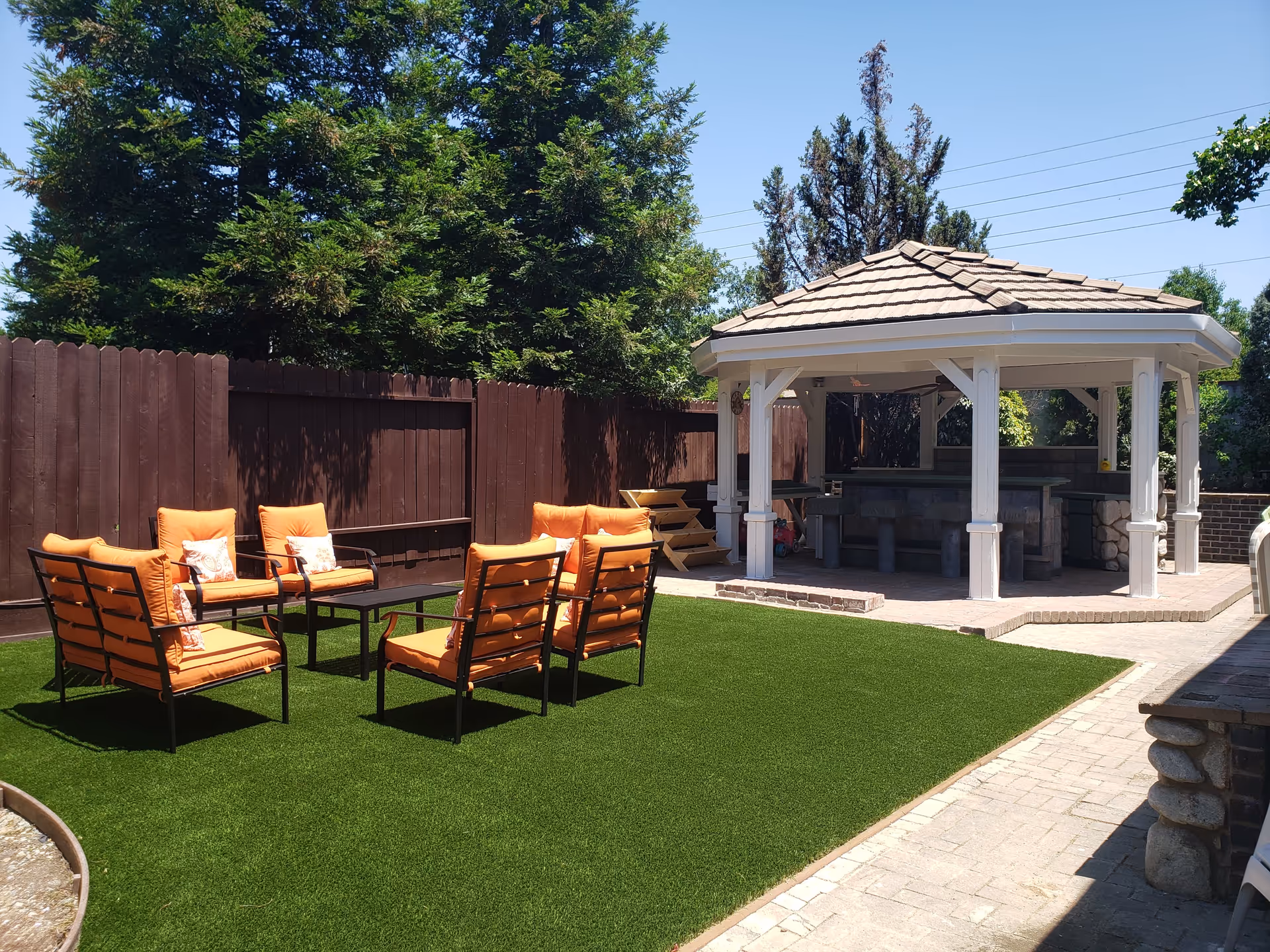 Outdoor seating area with six orange cushioned chairs arranged around a black coffee table on green artificial grass. In the background, there is a wooden gazebo with a tiled roof and bar-style seating underneath. The area is enclosed by a tall brown wooden fence and surrounded by trees under a clear blue sky.
