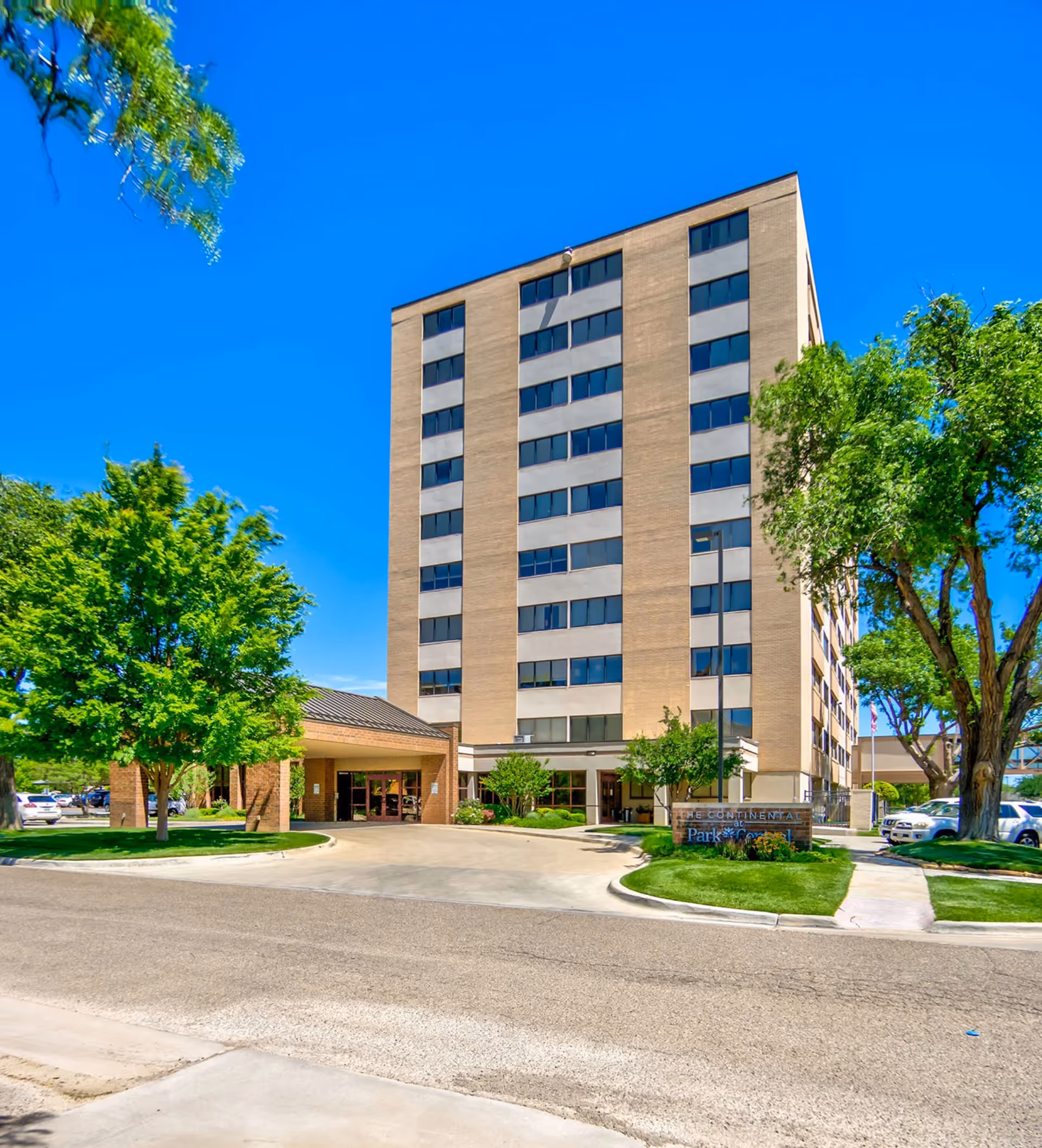 Front view of a tall beige senior living building with a main entrance and surrounding trees.