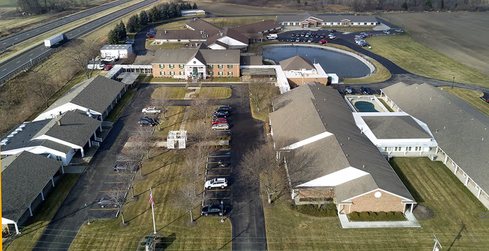 Aerial view of a senior living campus showing multiple single-story buildings, a parking lot, and a pond.
