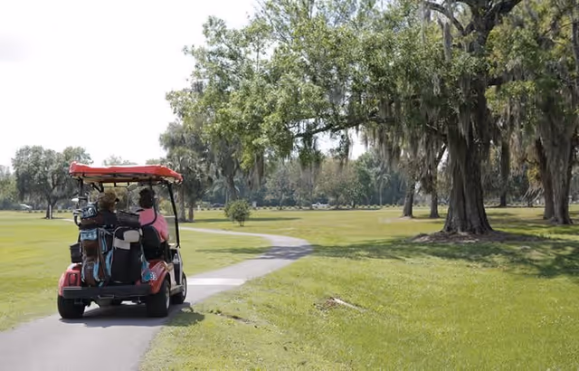 Two people riding in a red golf cart on a paved path through a green park-like area with large trees and grass.