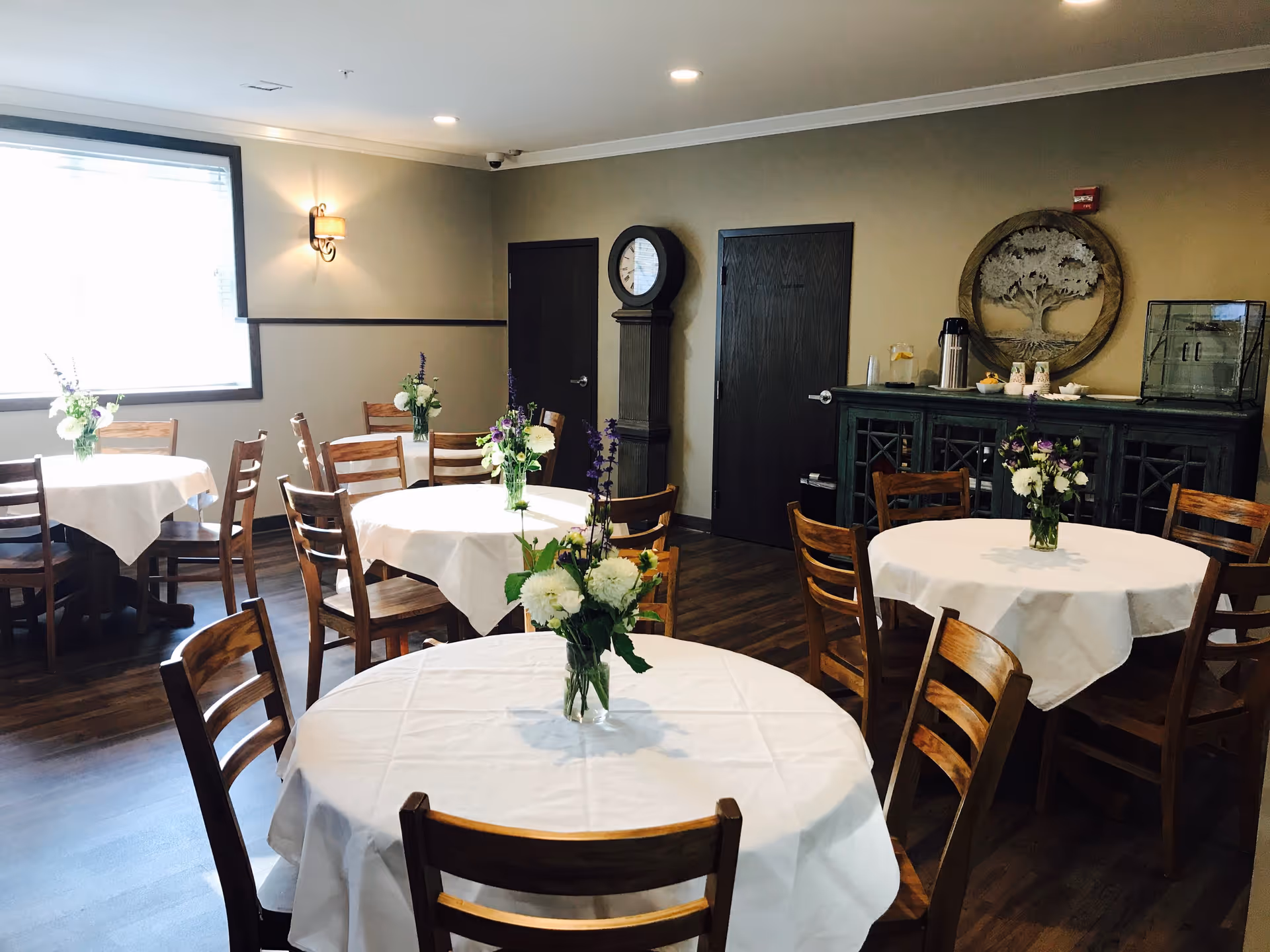 A dining room with several round tables covered with white tablecloths, each adorned with a vase of flowers. Wooden chairs surround the tables. The room has wooden flooring, a large window letting in natural light, a grandfather clock, and a dark wooden cabinet against the wall with a decorative tree artwork above it.