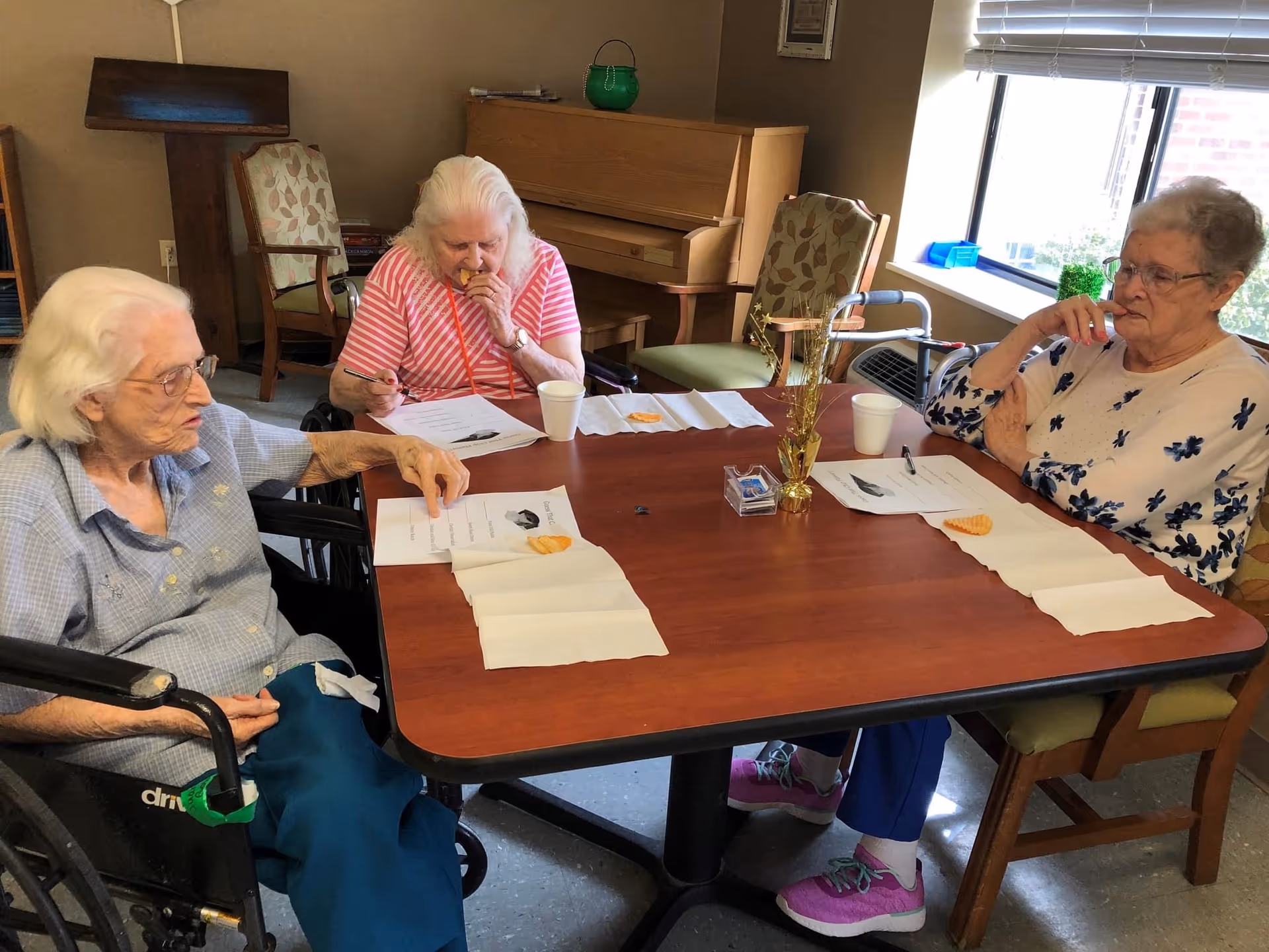Three elderly women seated around a table in a senior facility eating snacks and working on papers.