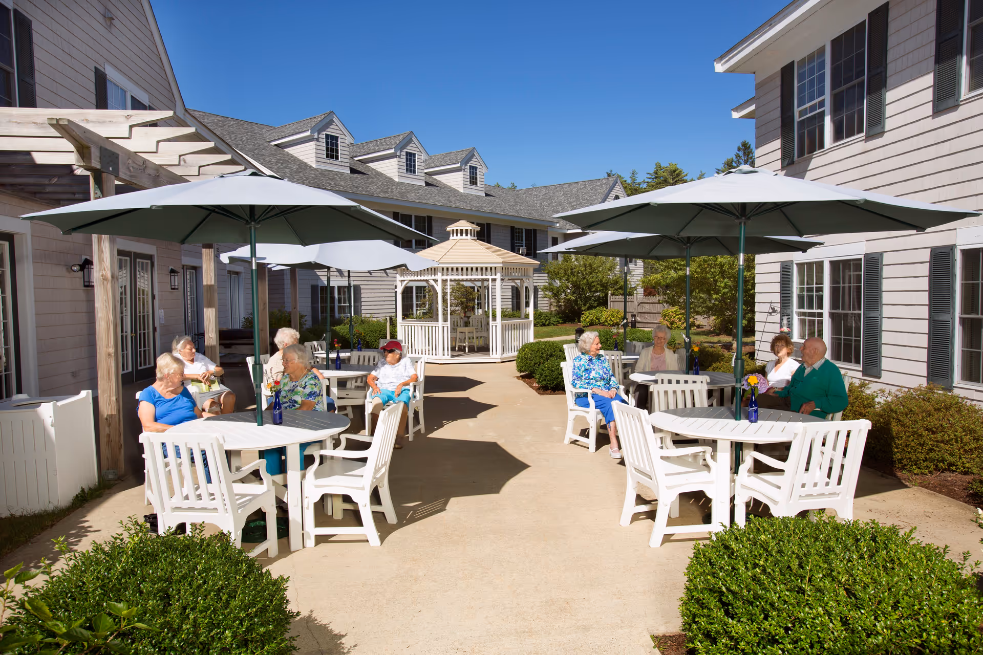 Outdoor patio area at Cape Cod Senior Residences with several elderly residents sitting at white tables under large umbrellas, surrounded by bushes and buildings with gray siding and multiple windows, and a white gazebo in the background under a clear blue sky.