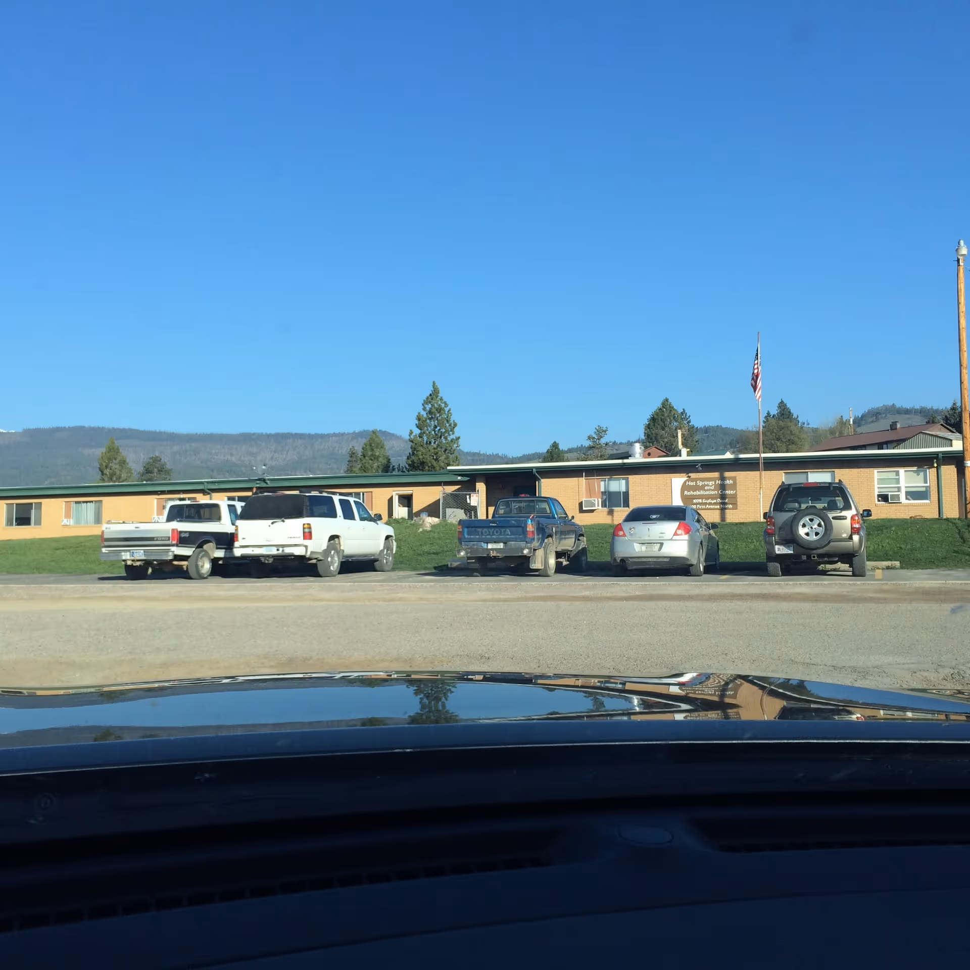 Exterior view of Hot Springs Health and Rehabilitation Center building with several parked vehicles in front, an American flag on a pole, and a clear blue sky with distant hills in the background.