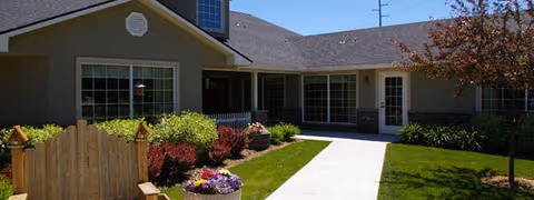 Front courtyard and entrance of a single-story assisted living building with a paved walkway, lawn, flower planters and a small wooden fence.