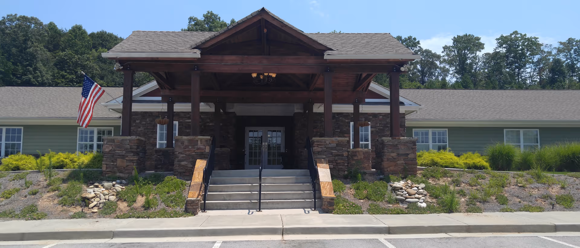 Front exterior view of a single-story building with a covered entrance supported by wooden beams and stone pillars. There are steps leading up to the entrance, an American flag on the left side, and landscaping with bushes and rocks in front of the building. Trees are visible in the background under a clear sky.