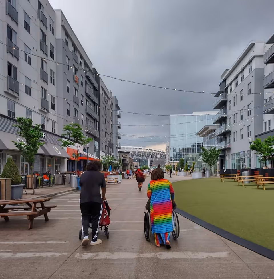 Outdoor plaza between modern buildings with string lights, seating, and people including wheelchairs.