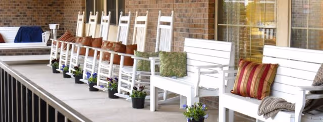 Row of white rocking chairs with cushions on a brick porch, potted plants nearby.