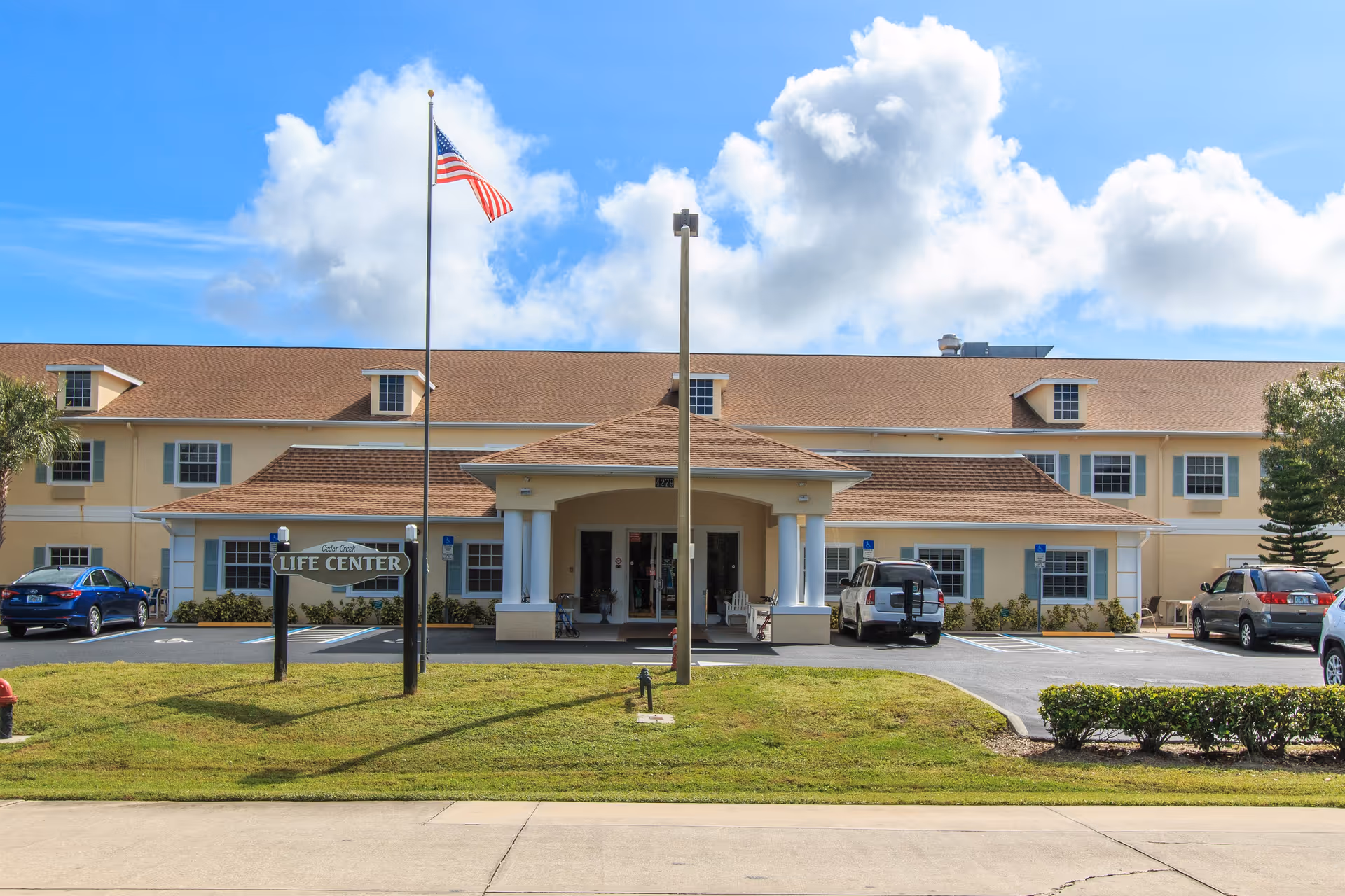 Front exterior view of Cedar Creek Assisted Living facility showing a two-story building with a brown roof, beige walls, and multiple windows. There is a covered entrance supported by white columns, an American flag on a flagpole, a sign that reads 'Cedar Creek Life Center,' and several parked cars in the parking lot. The sky is blue with some clouds.