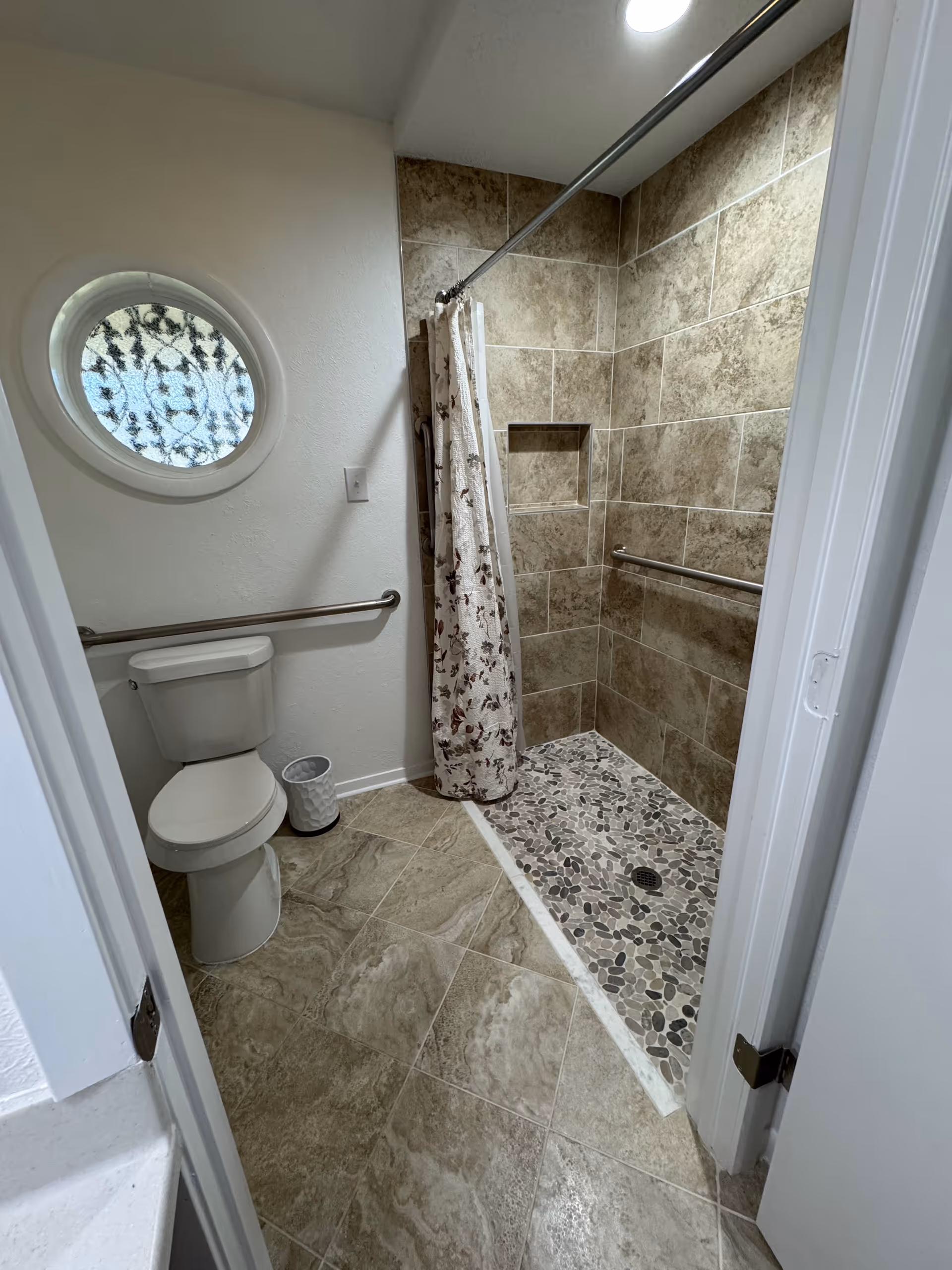 Compact bathroom with toilet, round window, and pebble-tile shower.