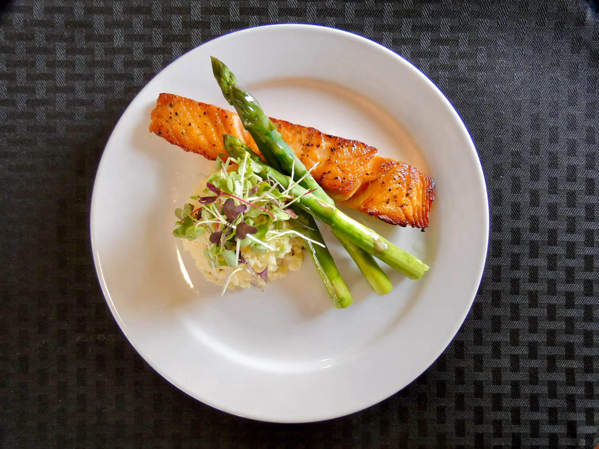 A plate featuring a portion of grilled salmon, accompanied by asparagus and a small serving of salad on a bed of rice, set against a textured black tablecloth.