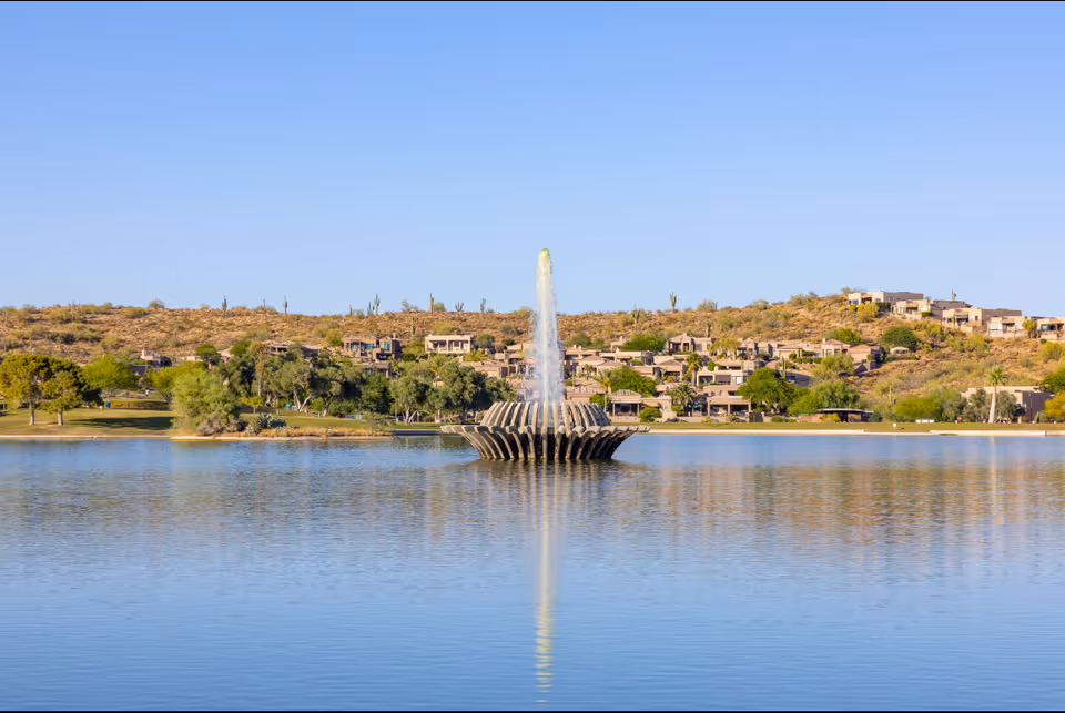 A tall central fountain in a lake with low hills and houses in the background.