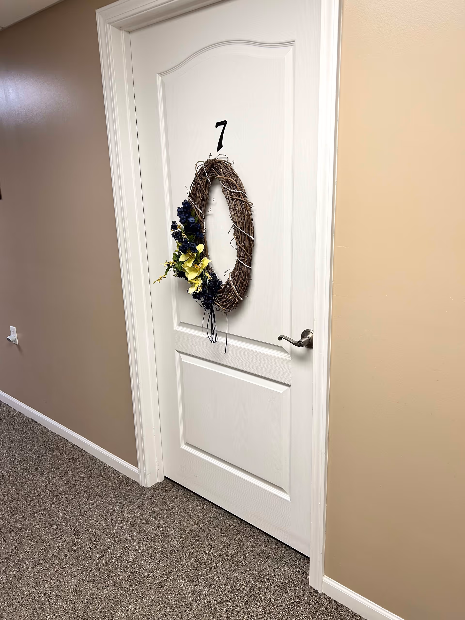 A white door in a hallway with the number 7 on it. The door has a decorative wreath made of twigs and adorned with yellow and dark blue flowers. The hallway walls are painted beige and the floor is carpeted in a neutral tone.