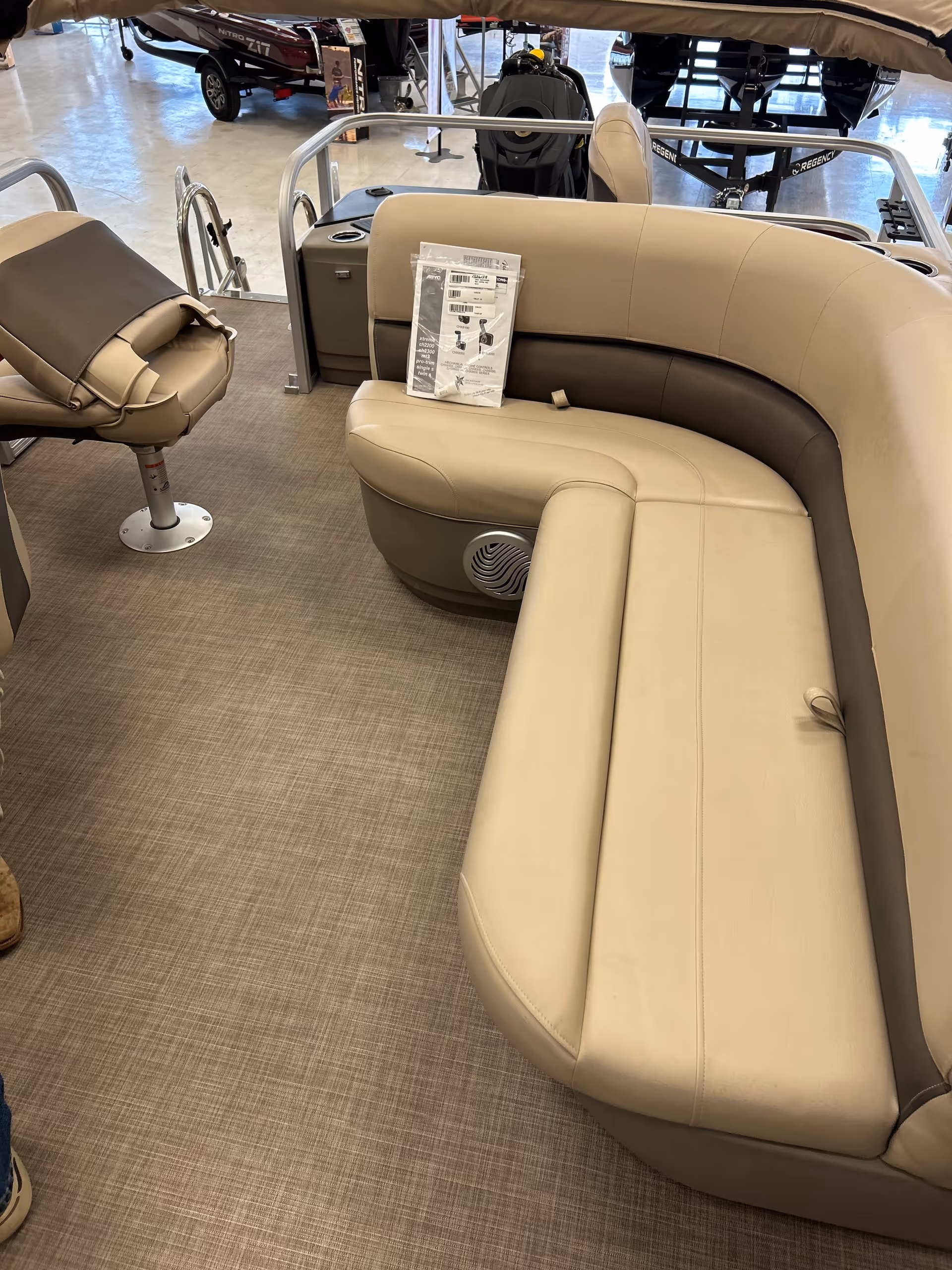 Interior seating area of a boat with beige and brown cushioned seats, a swivel chair, and a small speaker embedded in the seating. The floor has a textured, carpet-like surface. In the background, other boats and equipment are visible in a showroom setting.