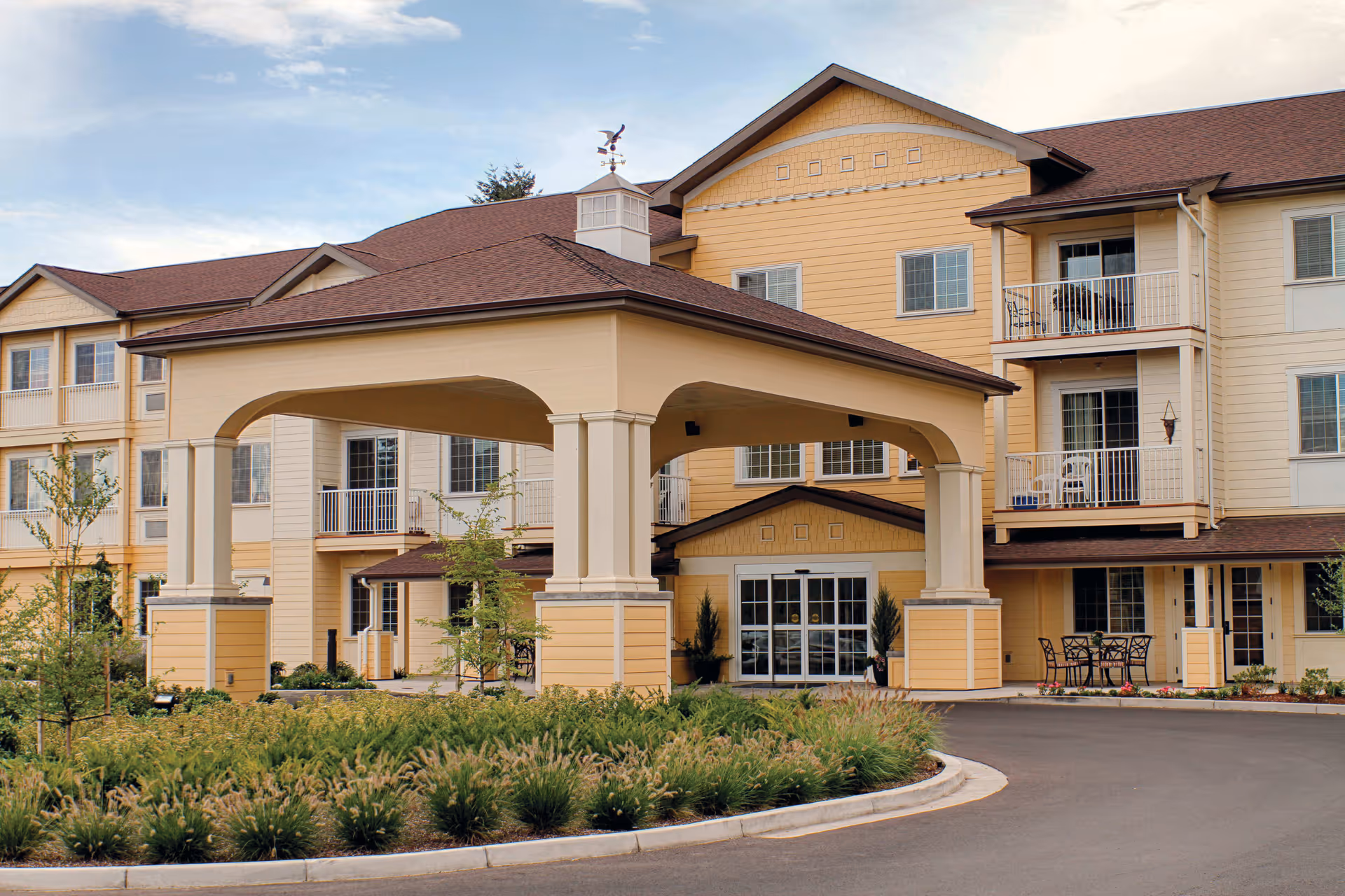 Exterior view of a senior living facility building with a covered entrance, multiple windows, balconies, and landscaped greenery in front.