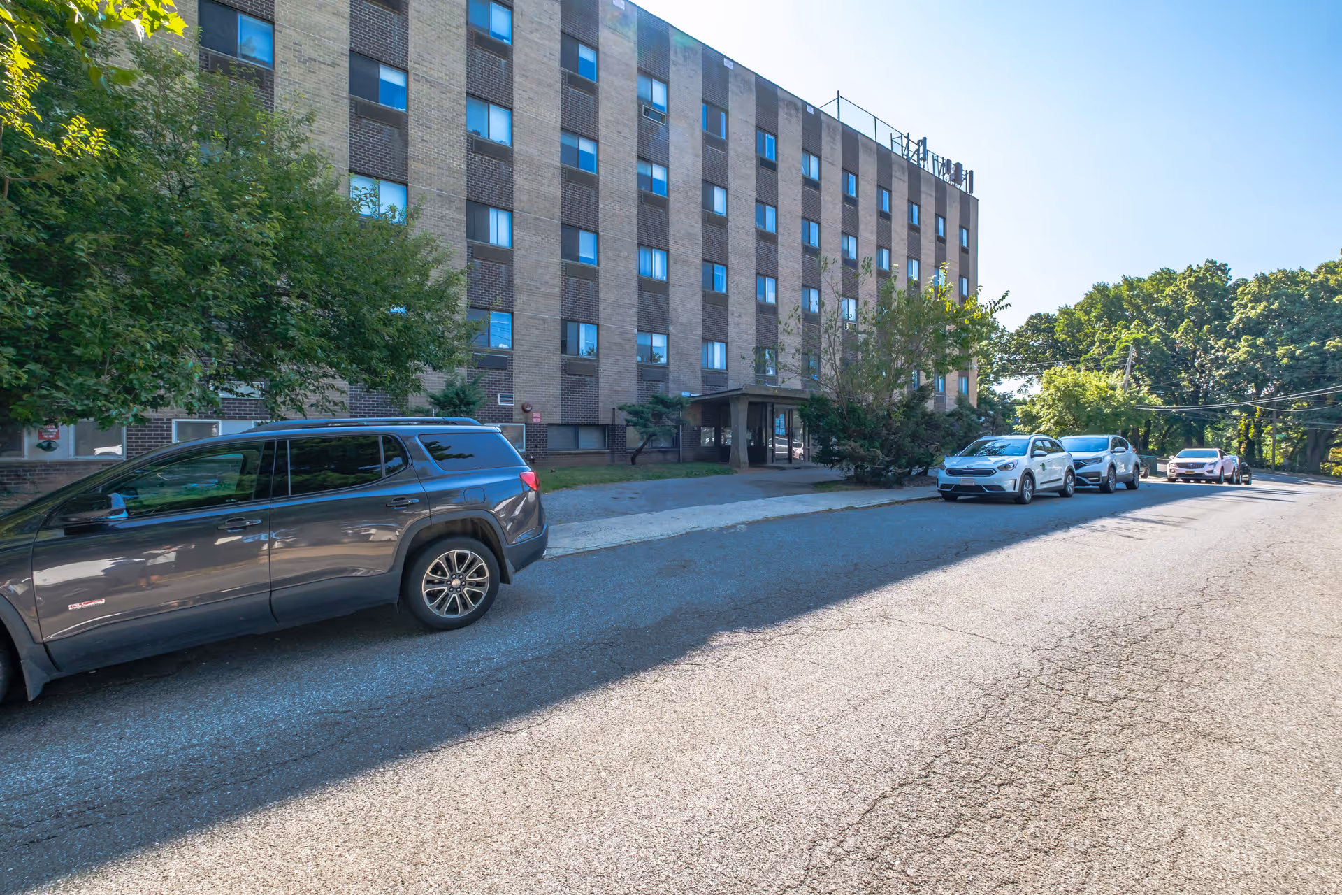 Multi-story brick residential building with a covered entrance, cars parked along the street, and trees lining the property.