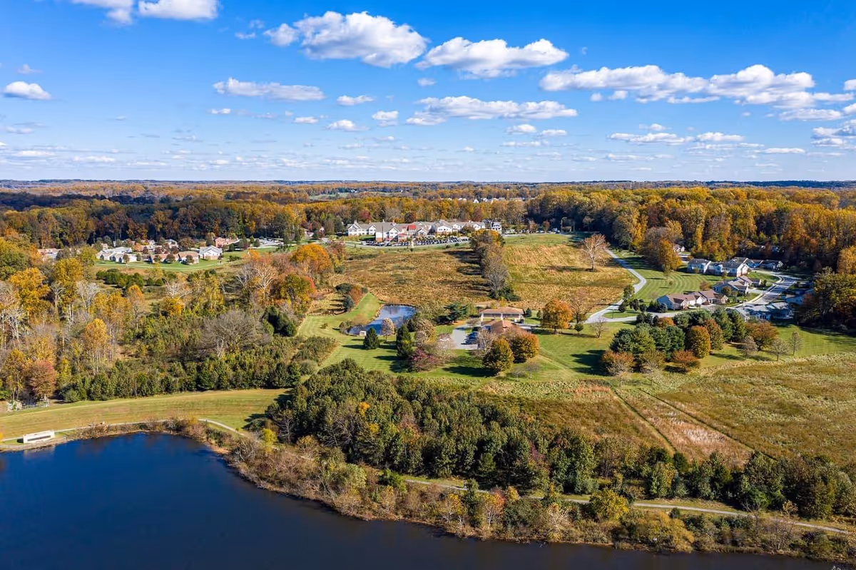 Aerial view of a large green landscape with a body of water in the foreground, surrounded by trees and open fields. In the distance, there are buildings and houses scattered among the trees under a partly cloudy blue sky.