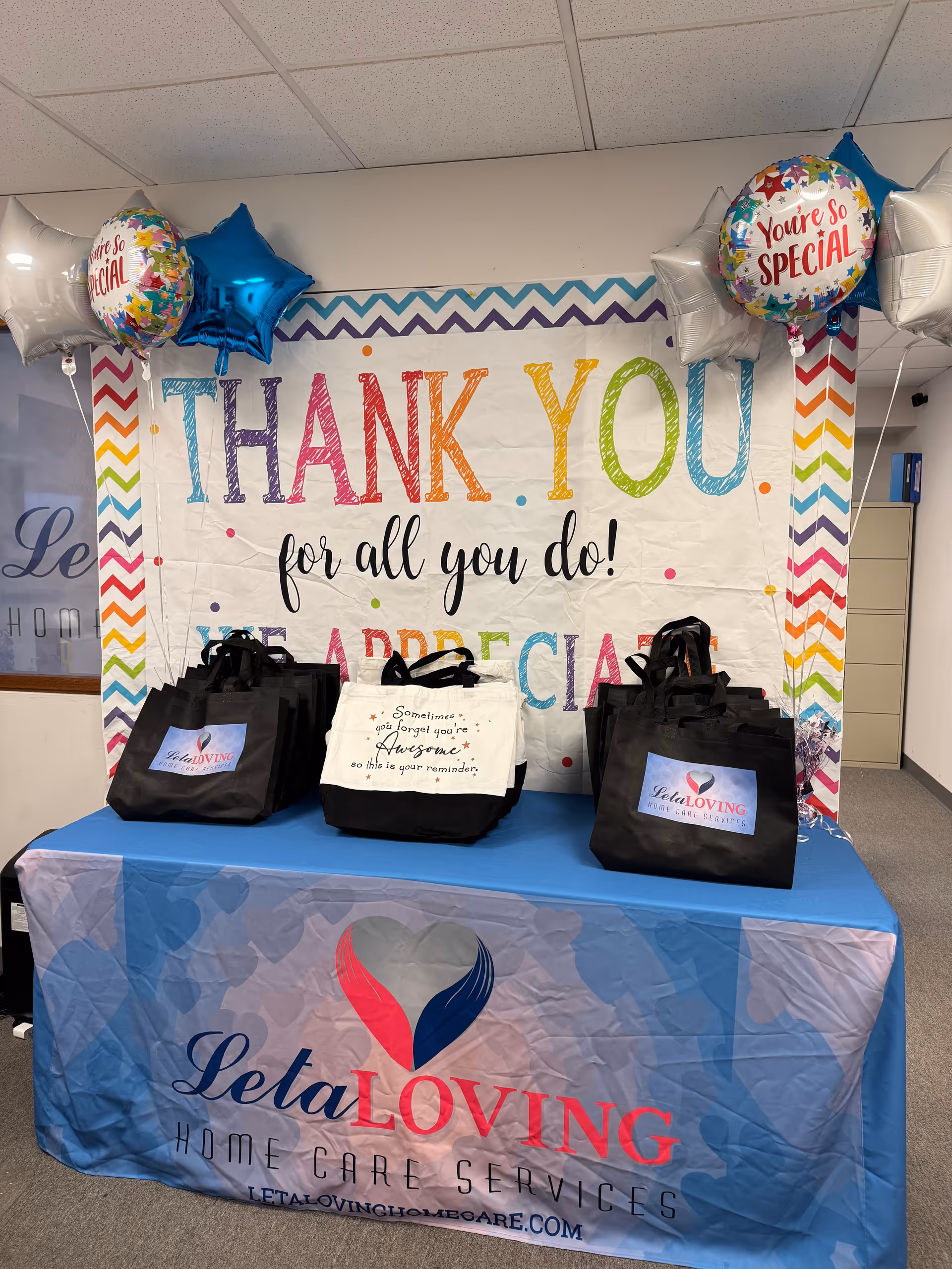 Indoor reception area with a colorful Thank You banner, balloons, and gift bags on a table.