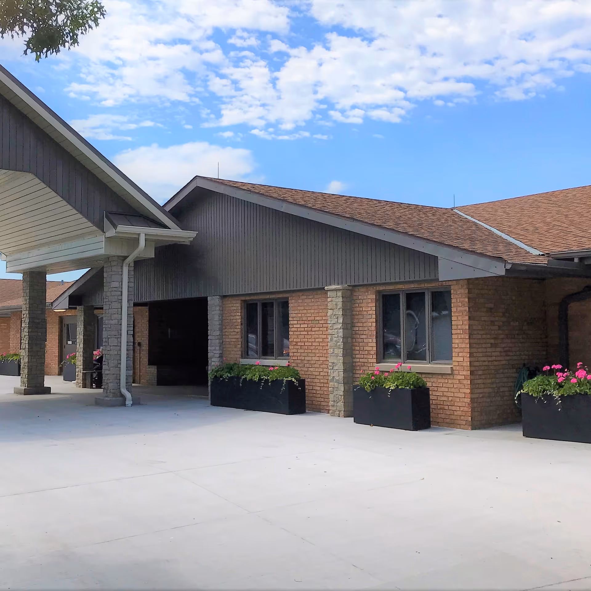 Brick senior living facility entrance with a covered porte-cochere, planter boxes with flowers, and a clear blue sky.