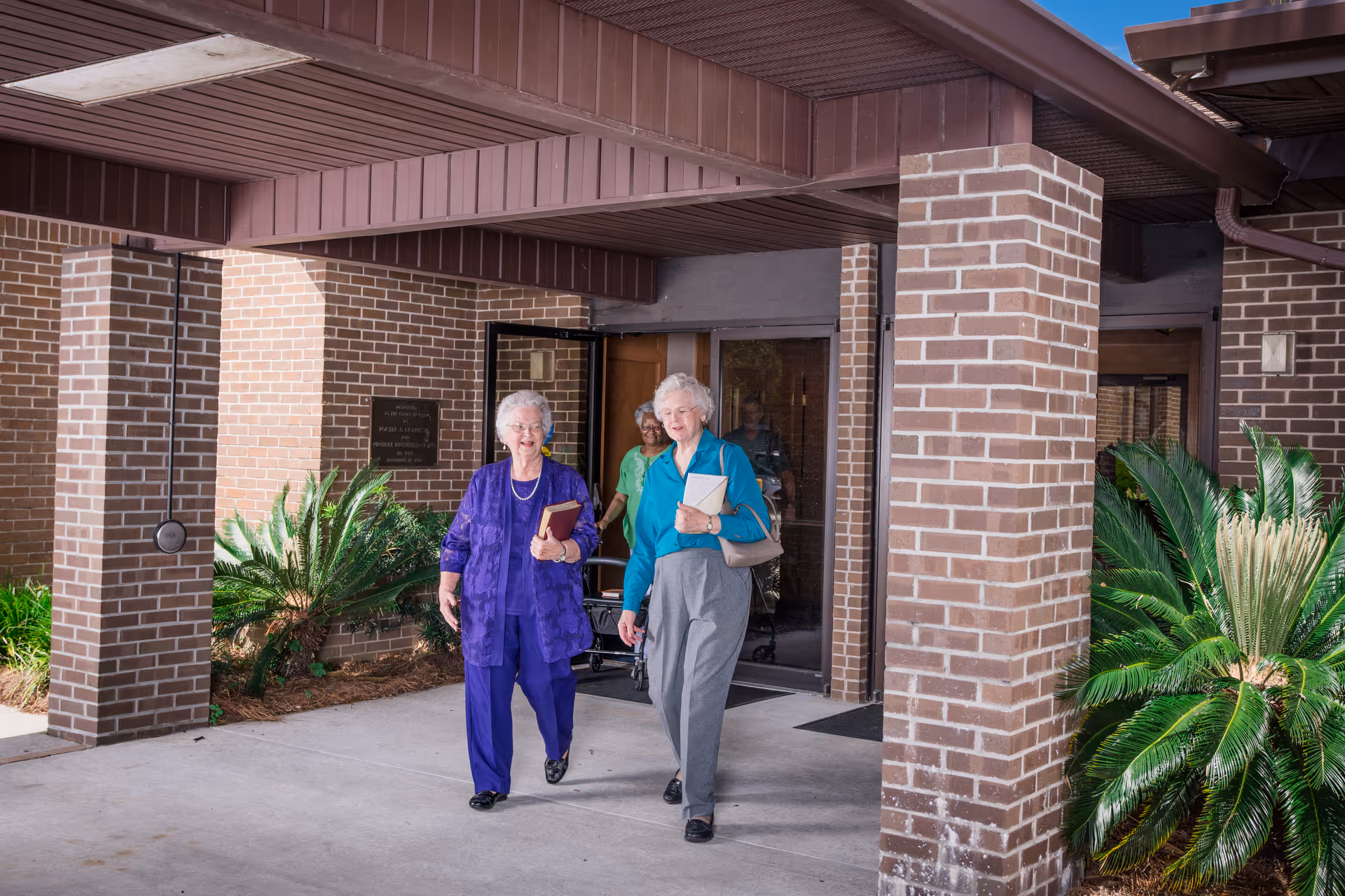 Two elderly women walking out of a brick building entrance under a covered portico, carrying books and a purse.