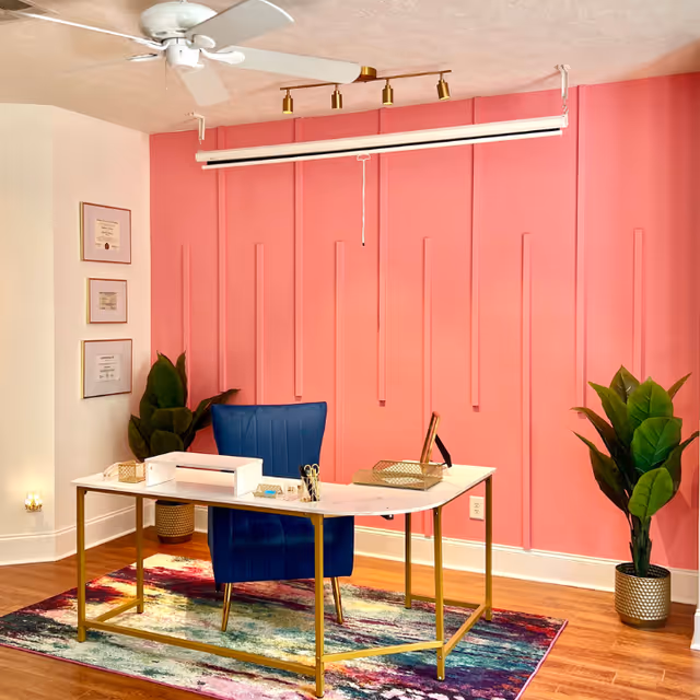 Pink-paneled office with white desk, blue chair, plants, and framed certificates.