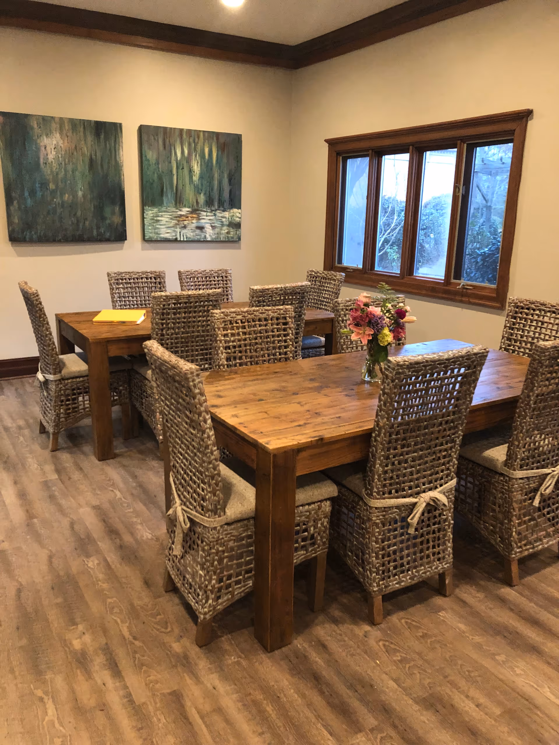 A cozy dining room with wooden tables, wicker chairs, wall art, and a flower centerpiece.