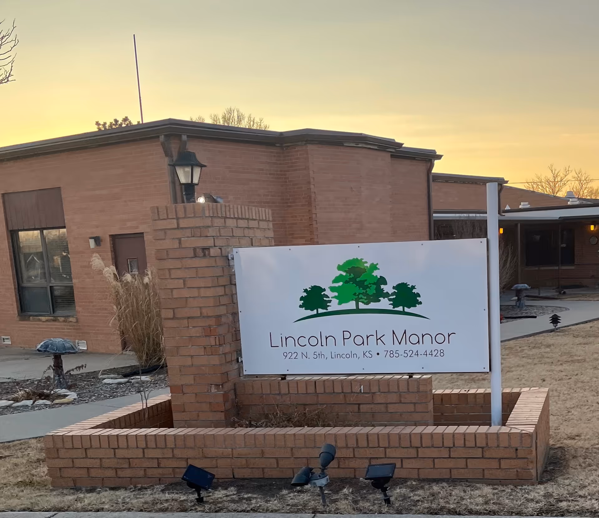 Front exterior of Lincoln Park Manor with a brick sign and landscaping at dusk.