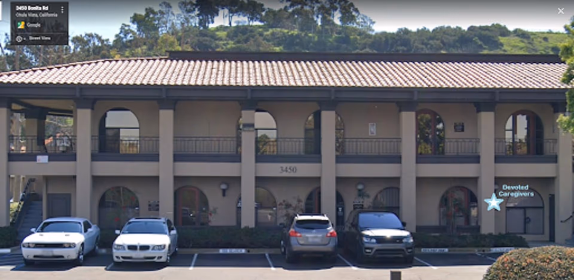 Two-story beige senior living facility named Devoted Caregivers with arches and parked cars in front.