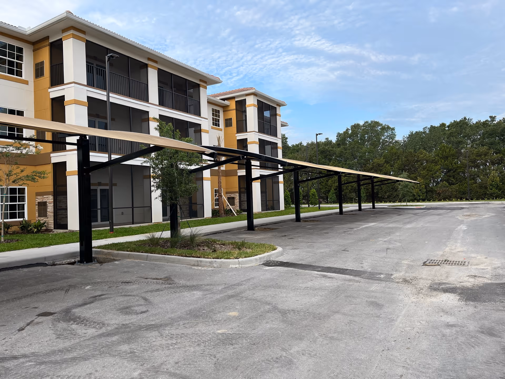Exterior view of a three-story residential building with screened balconies and long covered parking canopies in front.