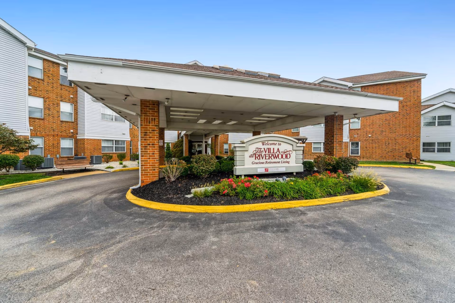 Covered drive-through entrance with a landscaped island and a sign reading "The Villa at Riverwood" in front of a multi-story brick senior living building.
