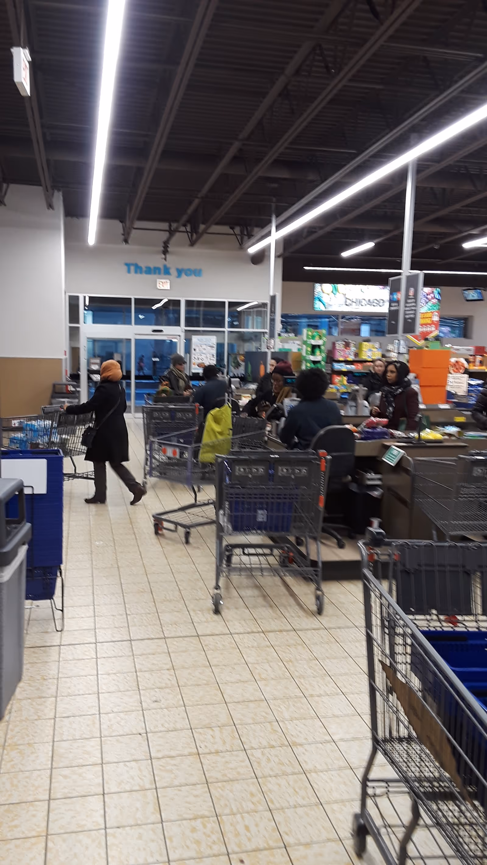 Shoppers push carts and wait at checkout in a bright supermarket interior.