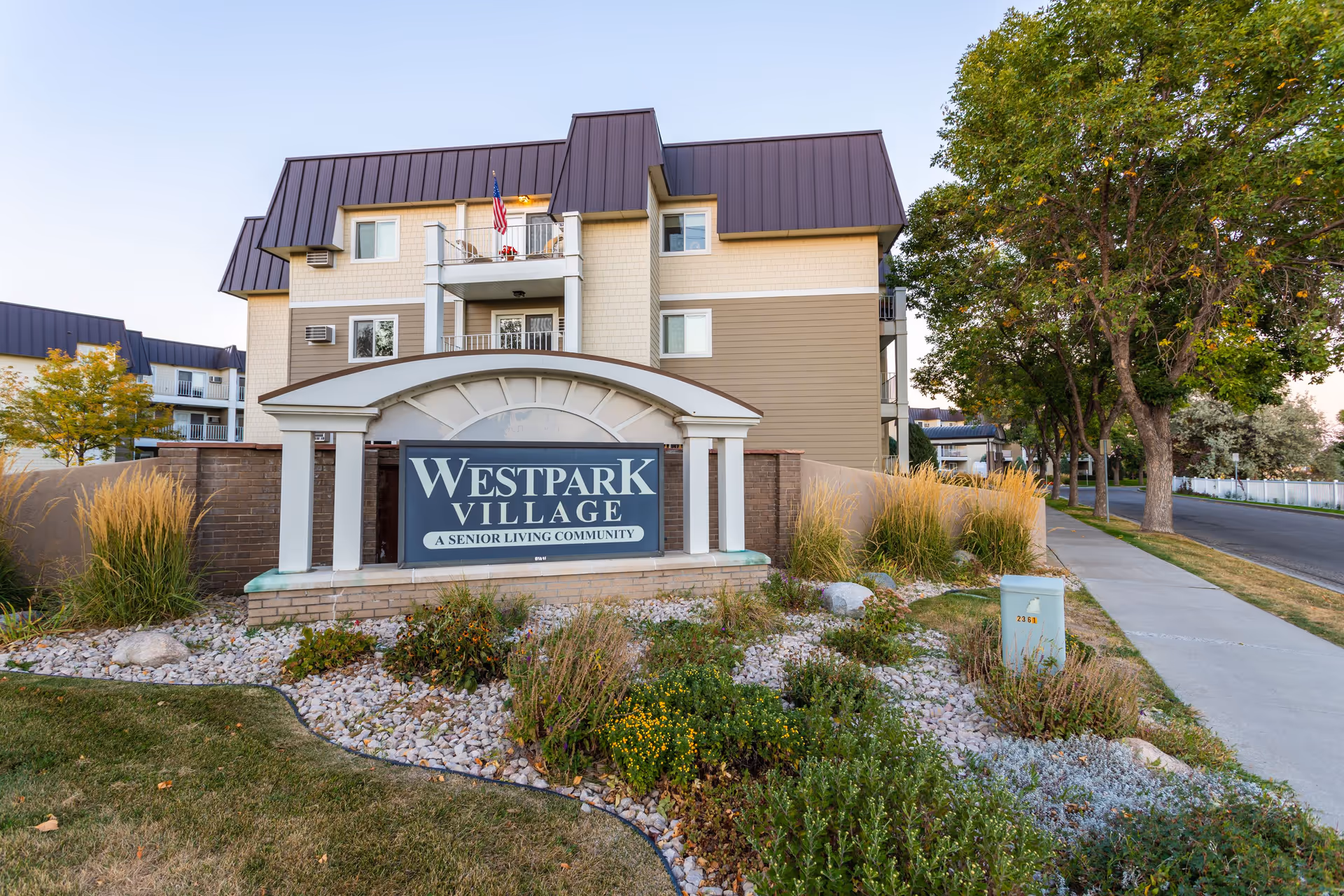 Exterior view of Westpark Village senior living community building with a landscaped garden and a sign in front displaying the facility name. The building has multiple floors with balconies and a brown roof. Trees and a sidewalk are visible along the street.