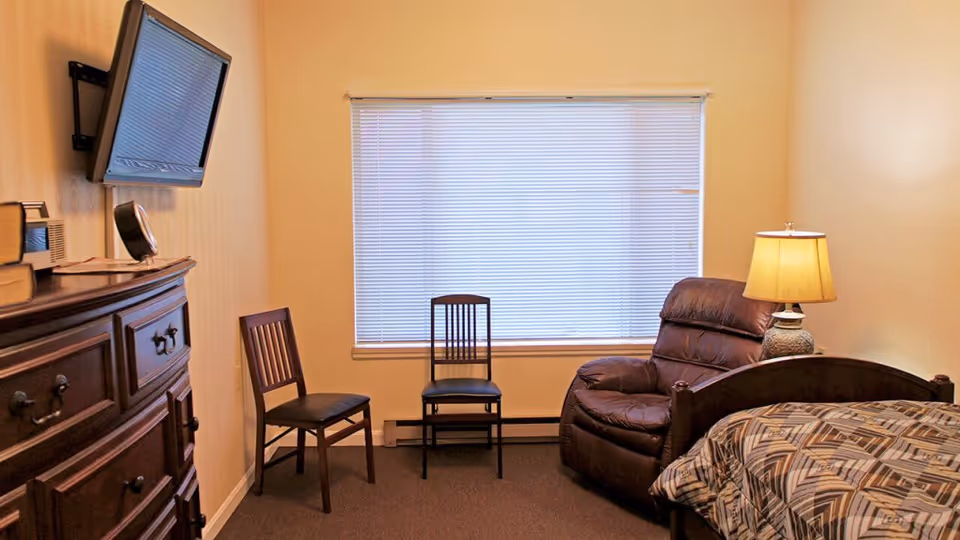 A small, cozy assisted living bedroom with a single bed covered in a patterned bedspread, a brown leather recliner chair, two wooden chairs, a wooden dresser with books and a clock on top, a wall-mounted flat-screen TV, and a window with closed blinds. A table lamp is placed on a small side table next to the recliner.