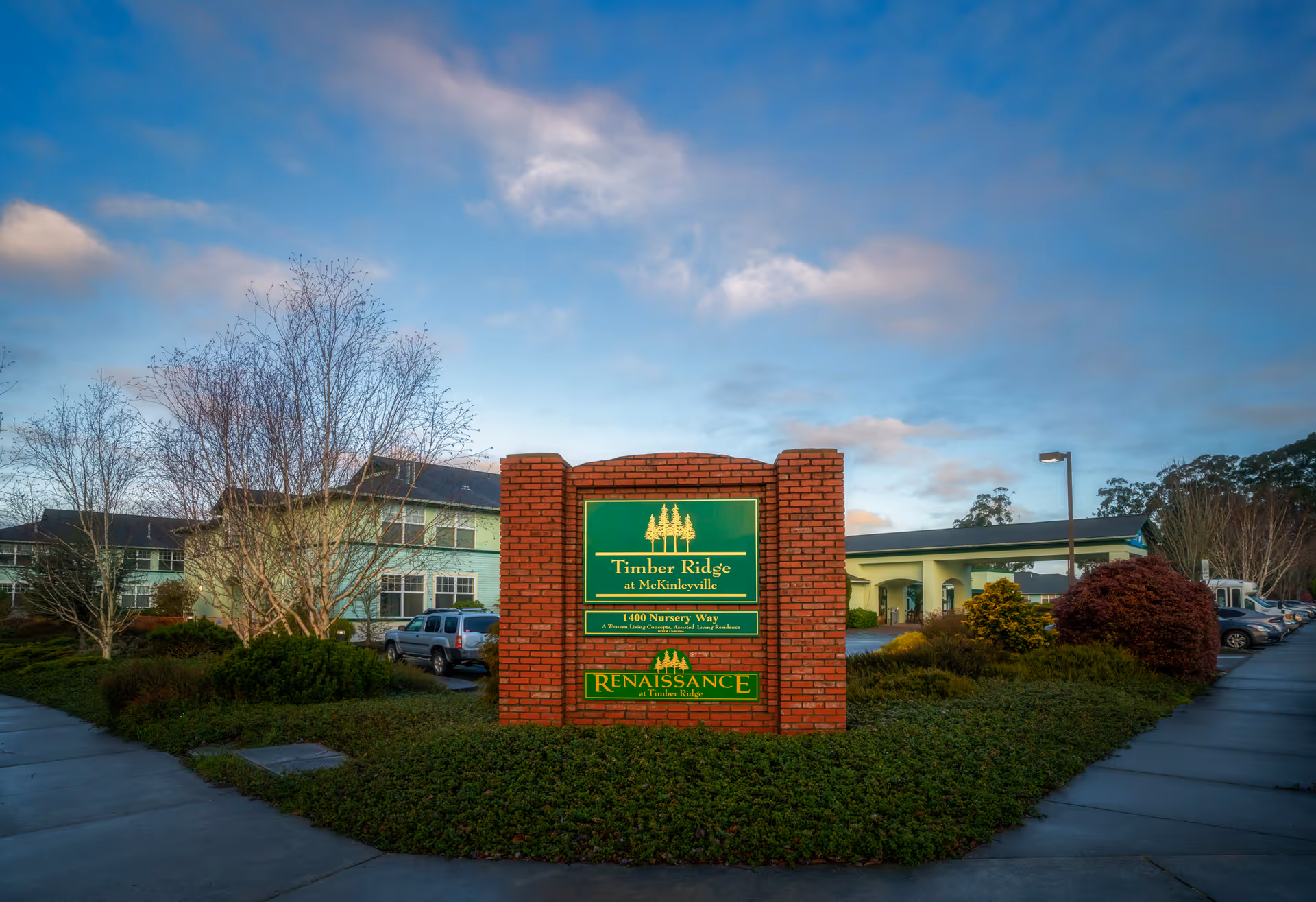 A brick entrance sign reading 'Timber Ridge at McKinleyville' set in landscaped grounds with the facility building and parked cars in the background under a blue sky.